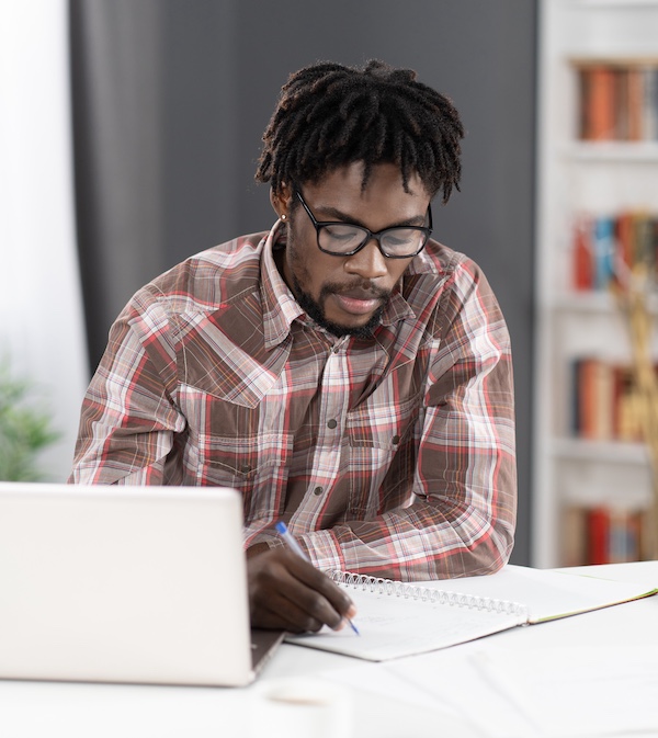 Young african student man making notes working at home working on laptop or listening teacher or boss during video call. African student study using laptop at home Young african student man making notes working at home working on laptop or listening teacher or boss during video call. African student study using laptop at home.